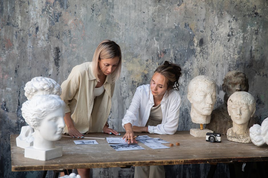 Two female artists discussing project ideas in a sculpture-filled art studio.